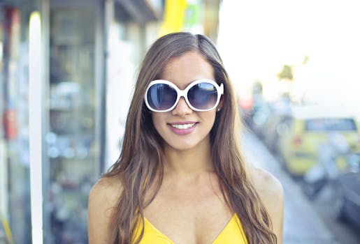 A stylish young woman with sunglasses smiles confidently on a sunny urban street.